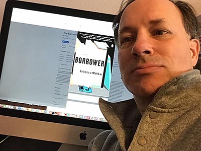 Preston in front of book club selection The Borrower: A Novel, book cover, on computer screen